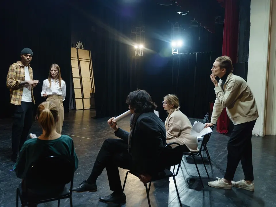 Grupa ljudi na probi na sceni, sjede u krugu i čitaju tekst